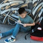 Child sitting on sidewalk writing in a notebook beside graffiti wall with a backpack.