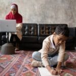 A Muslim woman in hijab working on a laptop while her son draws on the carpet.