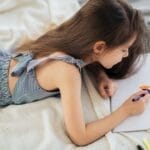 A young girl in a striped dress lying down, drawing with markers on white paper.