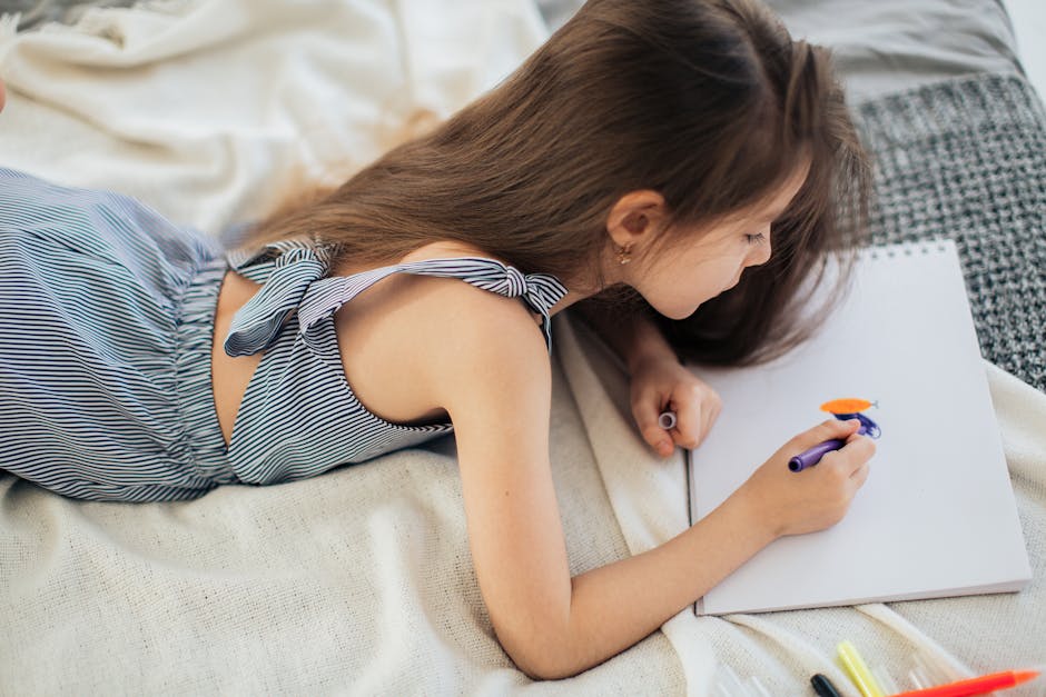 A young girl in a striped dress lying down, drawing with markers on white paper.