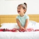 A happy child with Down syndrome smiling broadly while sitting on a bed indoors.