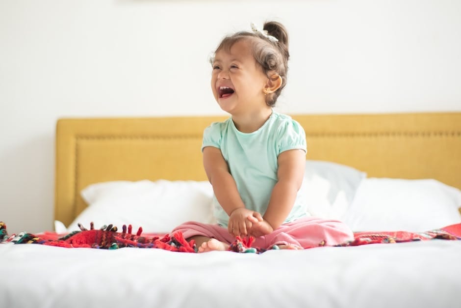 A happy child with Down syndrome smiling broadly while sitting on a bed indoors.