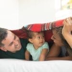 Joyful family of three sharing loving moments under a blanket indoors.