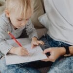 Young child focused on coloring paper with a red pencil, aided by an adult.