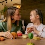 An adult woman and a child with Down syndrome engaging in playful interaction with wooden toys indoors.