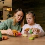 A mother and daughter engaging in play with wooden toys at a table indoors.