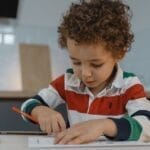 A young child focused on drawing with colored pencils at a table indoors, displaying creativity and concentration.