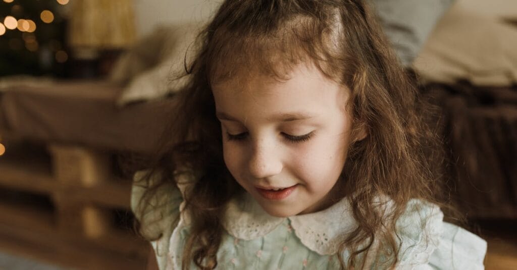 A young girl with long hair smiles gently in a cozy indoor setting.