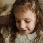 A young girl with long hair smiles gently in a cozy indoor setting.