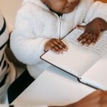 High angle of crop unrecognizable ethnic curious baby examining notepad near lady in casual outfit at table in bright apartment