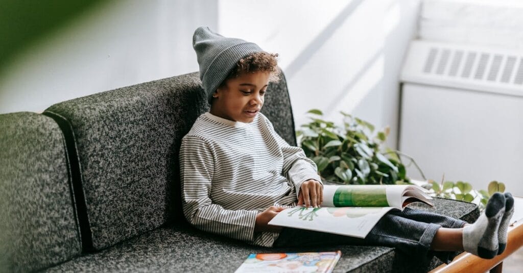 A young boy in a beanie and striped shirt reading a book on a sofa, surrounded by plants.