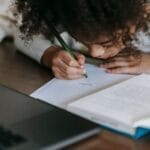 A young girl intently writes with a pencil, surrounded by educational materials at home.