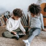 Thoughtful African American children in casual clothes sitting on floor against guitar and books and using tablet at home in daytime