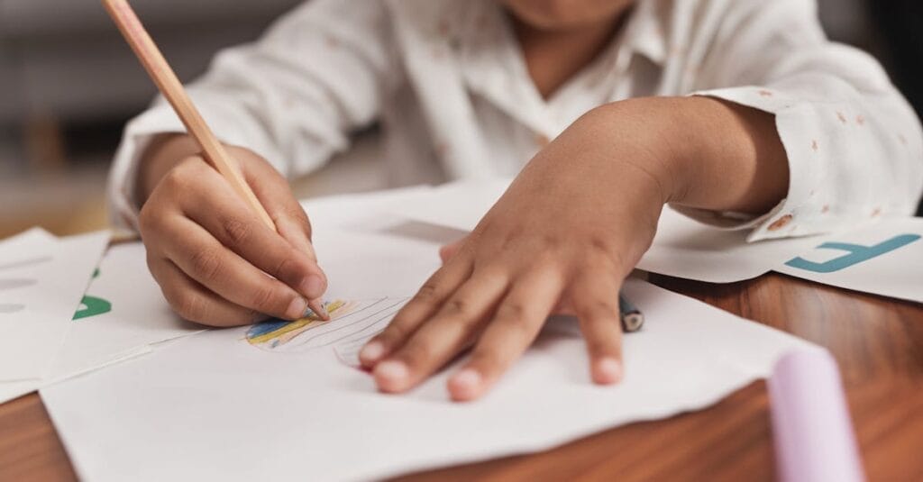 Child drawing with colored pencils on paper at a wooden table.