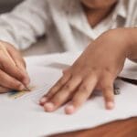 Child drawing with colored pencils on paper at a wooden table.