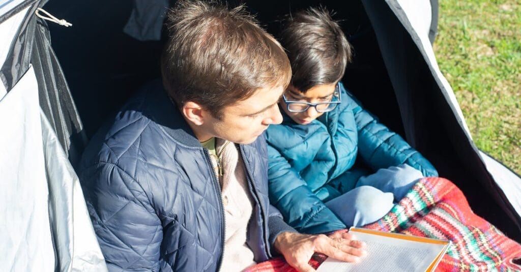 Father and son enjoy quality reading time in a cozy tent outdoors.