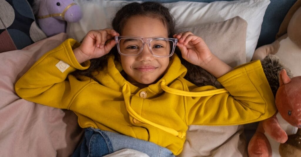 A young girl in glasses and a yellow hoodie smiling while lying on the bed with stuffed toys.