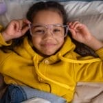 A young girl in glasses and a yellow hoodie smiling while lying on the bed with stuffed toys.