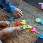 From above of crop anonymous ethnic baby and kid playing with assorted toy numbers and letters at wooden table in house