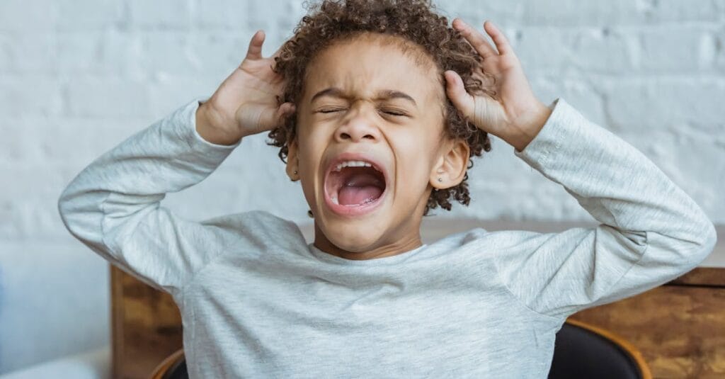 Young boy showing frustration indoors with hands on head, eyes closed, mouth open.