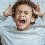 Young boy showing frustration indoors with hands on head, eyes closed, mouth open.