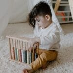 Young child sits on a carpet playing with a colorful wooden abacus in a cozy indoor setting.