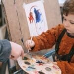 Young boy learning to paint with teacher's guidance during art class.