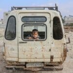 A young boy peers from an abandoned van in the desolate streets of Idlib, Syria, amidst war devastation.