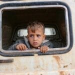 Scared ethnic child peeping out of aged transport hole with rust while looking away in daylight