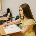 A young girl sitting alone in a classroom, studying and writing in a notebook.