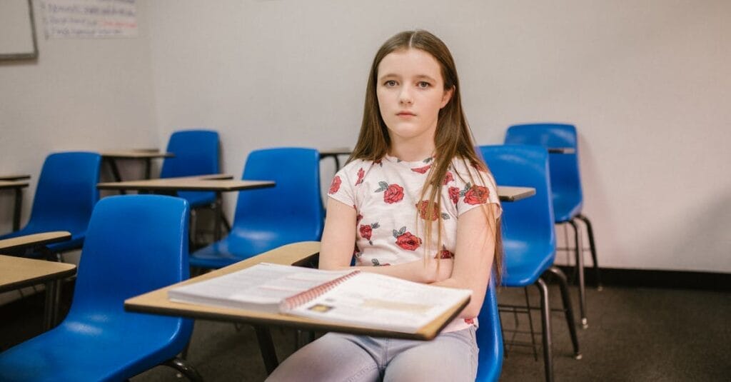 A young girl sits alone in an empty classroom looking pensive and isolated.