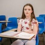 A young girl sits alone in an empty classroom looking pensive and isolated.