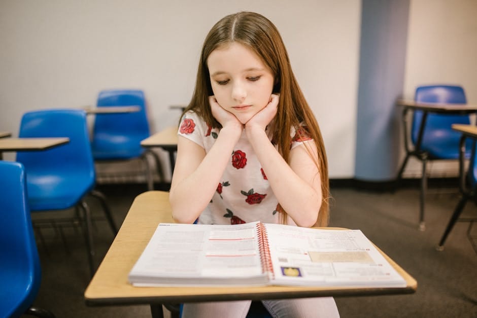 A young girl sits thoughtfully at her desk in a classroom, reading a textbook.