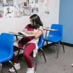 Young girl sitting alone in a classroom with blue chairs, symbolizing bullying and isolation.