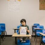 A child sits in an empty classroom, holding a sign against bullying.