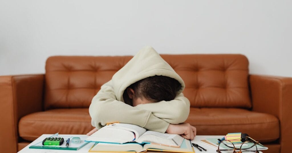 Child in hoodie leans over books, exhausted while studying on a couch.