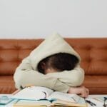 Child in hoodie leans over books, exhausted while studying on a couch.