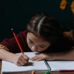 An East Asian child writing in a notebook at a wooden table, focused on homework indoors.