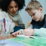 A young child writes while being supported by a teacher, focusing on learning.