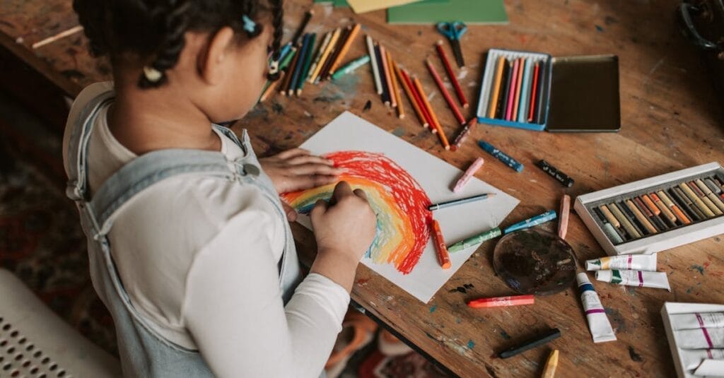 Young child creating a vibrant rainbow drawing at a wooden table with various art supplies.