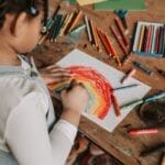 Young child creating a vibrant rainbow drawing at a wooden table with various art supplies.