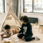 High angle of African American mother sitting on floor near girl reading book near play tent in studio