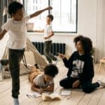 Full body of African American mother sitting on wooden floor near daughter writing in copybook in light room with playing boys