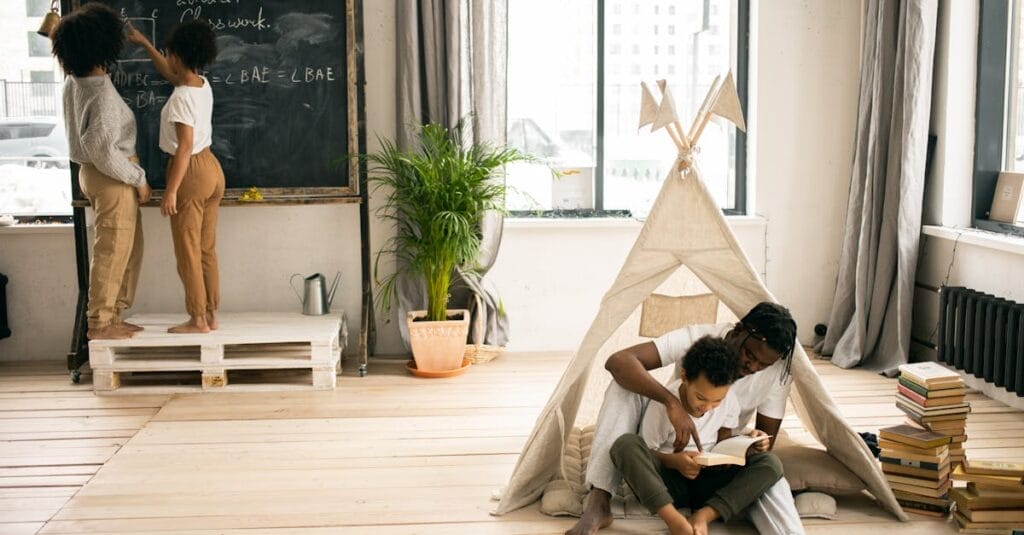 Full body of young African American man sitting on floor and helping little son to read book near wife and daughter writing on blackboard while doing homework assignment