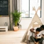 Full body of young African American man sitting on floor and helping little son to read book near wife and daughter writing on blackboard while doing homework assignment