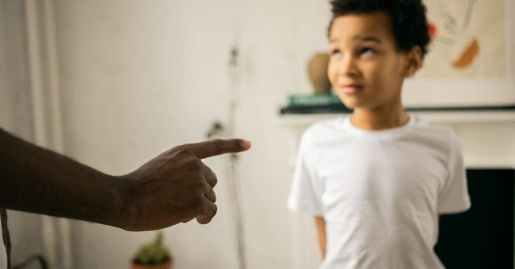 A father points and scolds his son indoors, emphasizing discipline.