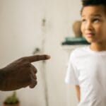 A father points and scolds his son indoors, emphasizing discipline.