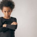 Portrait of a young child with crossed arms and a serious expression in a studio setting.