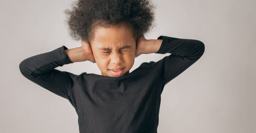 Unhappy African American girl with Afro hairstyle covering ears while standing on white background with closed eyes in light studio