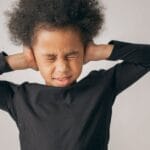 Unhappy African American girl with Afro hairstyle covering ears while standing on white background with closed eyes in light studio